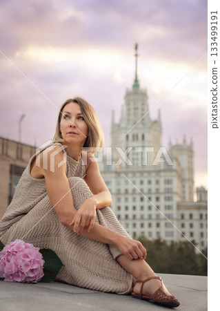 A woman sits at sunset with a pink hydrangea placed beside her, framed by the Kotelnicheskaya Embankment Building and soft pastel evening clouds A woman sits at sunset with a pink hydrangea placed beside her, framed by the Kotelnicheskaya Embankment Building and soft pastel evening clouds 133499191