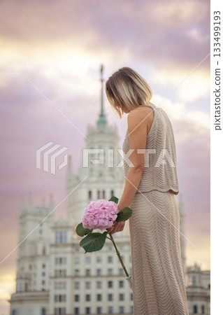 A woman stands at sunset holding a pink hydrangea, with the Kotelnicheskaya Embankment Building rising behind her in warm pastel light A woman stands at sunset holding a pink hydrangea, with the Kotelnicheskaya Embankment Building rising behind her in warm pastel light 133499193
