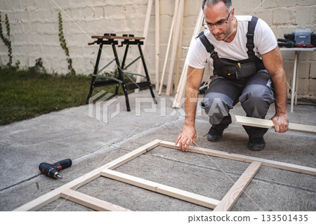 Mature male carpenter using electric screwdriver on a wooden plank outdoors 133501435