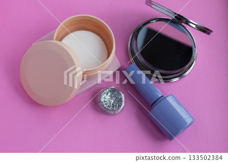 Cosmetics arrangement on a vibrant pink background featuring a compact mirror, loose glitter, a container of nail polish remover wipes, and nail polish, symbolizing beauty and self care 133502384