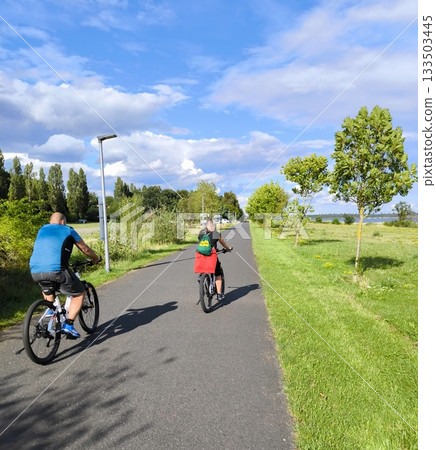 Two cyclists ride along a scenic road along a lake on a sunny day 133503445