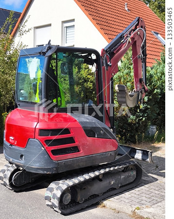 A compact mini excavator on a crawler track against the backdrop of a private residential building A compact mini excavator on a crawler track against the backdrop of a private residential building 133503465