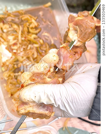 Preparing for the barbecue, a glove sleeve threads raw marinated meat onto a kebab skewer. 133503469