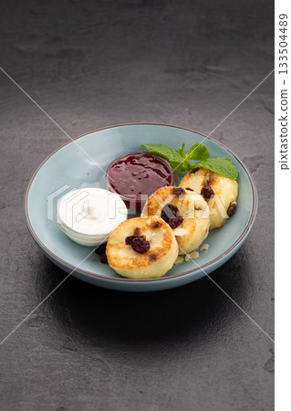 Syrniki with sour cream and berry jam in a blue bowl on dark stone background 133504489