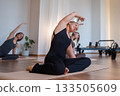 Group of woman carefully doing mermaid exercise, woman engaging core during seated twist in bright studio 133505609