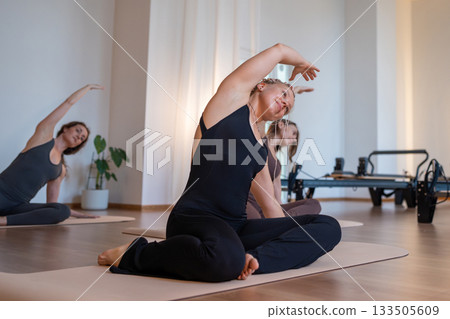 Group of woman carefully doing mermaid exercise, woman engaging core during seated twist in bright studio 133505609