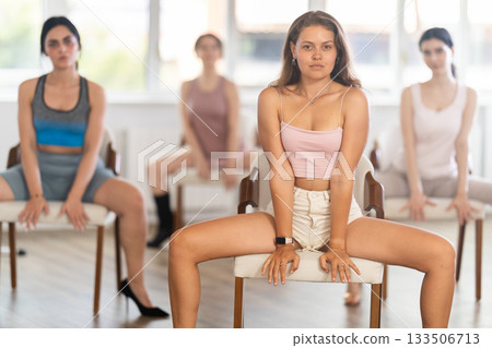Slender athletic young women perform various dance movements with legs while sitting on chairs 133506713