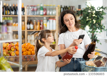 Girl with mother customers at shop scans QR code on juice package using phone camera Girl with mother customers at shop scans QR code on juice package using phone camera 133506803