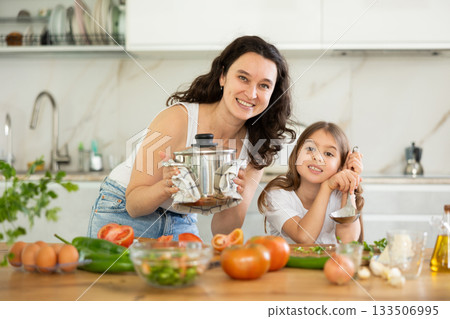 Mother holding saucepan and preteen daughter standing at table 133506995