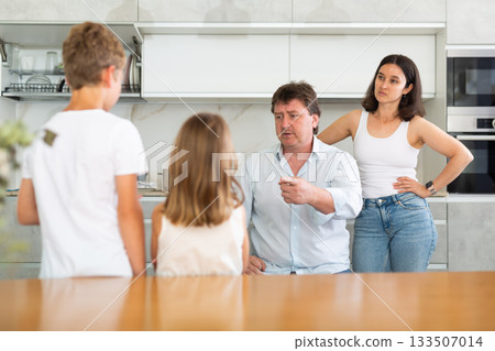 At home in kitchen, outraged mom and dad scold their son and daughter for disobedience. At home in kitchen, outraged mom and dad scold their son and daughter for disobedience. 133507014