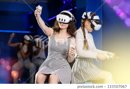 Girls wearing virtual reality glasses play in pairs, against each other, manipulating objects or shooting at targets using joysticks in their hands 133507067