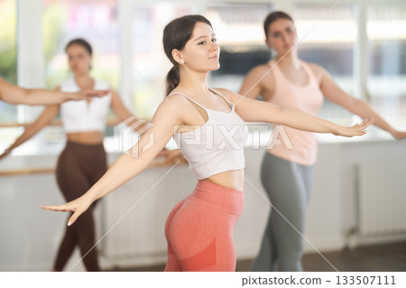 Pretty young girl ballerina practicing arabesque pose with group of dancers in dancing hall Pretty young girl ballerina practicing arabesque pose with group of dancers in dancing hall 133507111