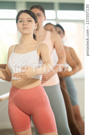 Happy young girl in sportswear, participating first position of ballet stance at barre with group youth people standing in row in choreography studio Happy young girl in sportswear, participating first position of ballet stance at barre with group youth people standing in row in choreography studio 133507130