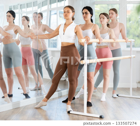 Group of happy fit young girls standing in row together while doing ballet dance moves dancers standing with one hand on barre and reaching over hand in ballet room 133507274