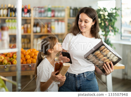 Daughter and mother choose delicious cookies in supermarket - scan QR code on label with smartphone Daughter and mother choose delicious cookies in supermarket - scan QR code on label with smartphone 133507948