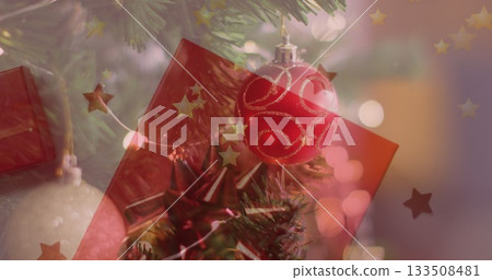 Shimmering red Christmas bauble with gold glitter on pine branch in living room, with star garland Shimmering red Christmas bauble with gold glitter on pine branch in living room, with star garland 133508481