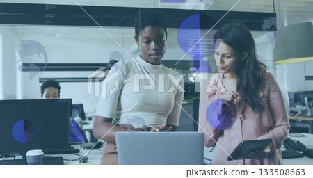 Two coworkers wearing cream top and pink blouse discussing data at laptop workstation, with tablet Two coworkers wearing cream top and pink blouse discussing data at laptop workstation, with tablet 133508663