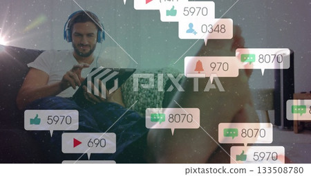 Sitting adult male wearing headphones tapping tablet in lounge, with floating notification icons Sitting adult male wearing headphones tapping tablet in lounge, with floating notification icons 133508780