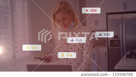 Using smartphone and laptop, woman wearing polka-dot blouse in kitchen with notification icons 133508789