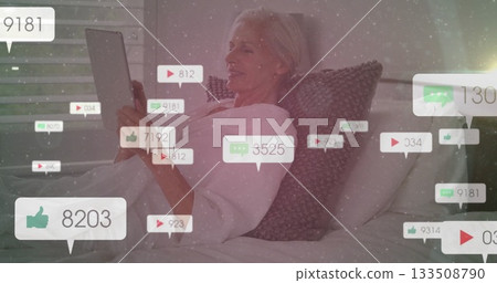 Reclining senior woman holding tablet computer in home bedroom, with floating social media icons 133508790