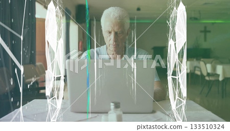 Working senior man focusing on silver laptop at church hall table, with shakers and overlays 133510324