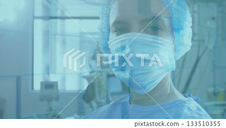 Standing surgeon in blue gown, mask and bouffant cap behind glass in operating room, with monitors Standing surgeon in blue gown, mask and bouffant cap behind glass in operating room, with monitors 133510355