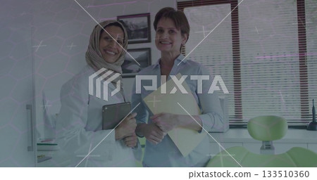 Standing women wearing lab coat and shirt holding tablet and folder in practice, with dental chair 133510360