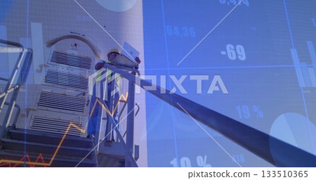 Engineer wearing safety helmet and vest holding laptop beside tank, with financial graph overlay 133510365