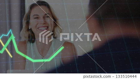 Smiling adult woman in white blouse reviewing data at office desk, with digital line graph overlay Smiling adult woman in white blouse reviewing data at office desk, with digital line graph overlay 133510444