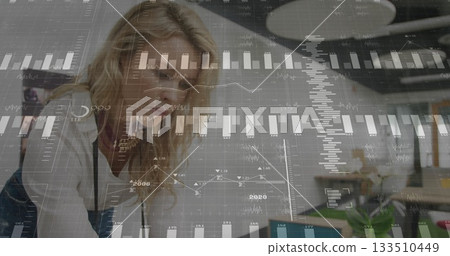 Leaning data analyst reviewing charts on laptop at open plan office desk, with notebook, plant 133510449