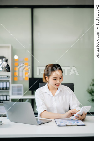 Asian woman with a smile standing holding notepad and tablet at the office. Asian woman with a smile standing holding notepad and tablet at the office. 133511342