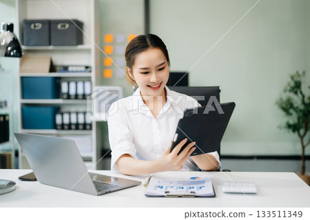 Asian woman with a smile standing holding notepad and tablet at the office. 133511349