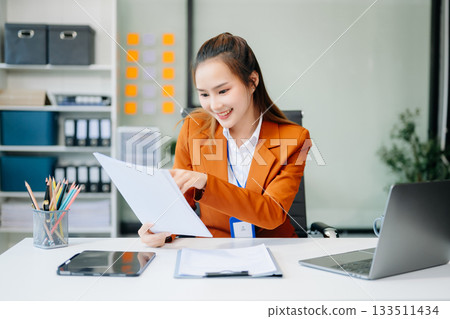 Asian businesswoman working with working notepad, tablet and laptop documents talking on the smartphone, tablet and laptop video call tax 133511434