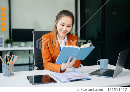 Asian woman with a smile standing holding notepad and tablet at the office. Asian woman with a smile standing holding notepad and tablet at the office. 133511437