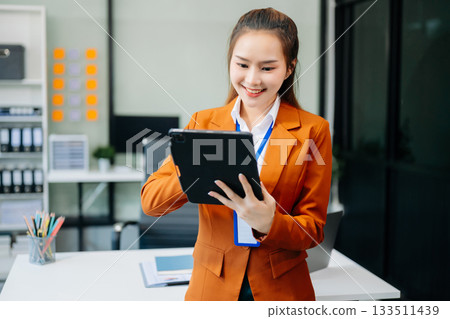 Asian woman with a smile standing holding notepad and tablet at the office. 133511439