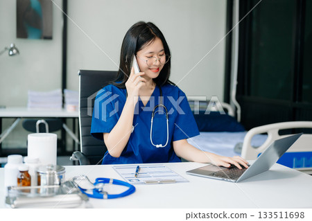 Confident young Asian female doctor in white medical uniform sit at desk working on computer. Smiling use laptop write in medical journal in clinic Confident young Asian female doctor in white medical uniform sit at desk working on computer. Smiling use laptop write in medical journal in clinic 133511698