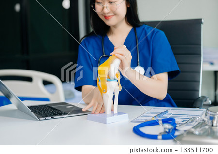 Confident young Asian female doctor in white medical uniform sit at desk working on computer. Smiling use laptop write in medical journal in clinic 133511700