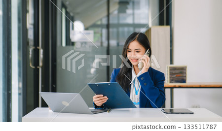 Young Asian businesswoman working with working notepad, tablet and laptop documents talking on the smartphone, tablet and laptop video call Young Asian businesswoman working with working notepad, tablet and laptop documents talking on the smartphone, tablet and laptop video call 133511787