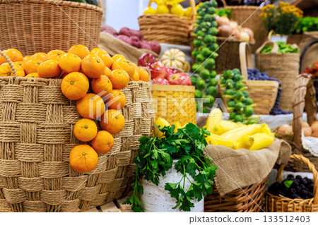Bright oranges hang from basket among various fruits vegetables in lively market setting. 133512403