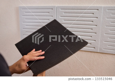 Closeup of foam insulation method, Hand pressing textured acoustic foam onto wall surface, Detailed shot of fingers securing black foam panels on gray wall Comparison of different foam rubber and 133515046