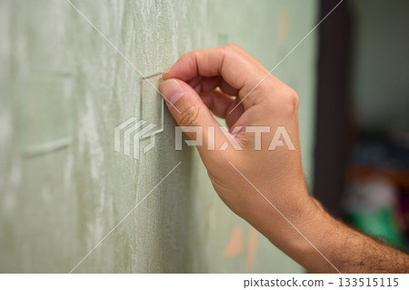 Closeup of textured plaster wall prep, Gently detaching thin tape from wall surface, Homeowner carefully removing adhesive strip to ready wall for repainting process 133515115