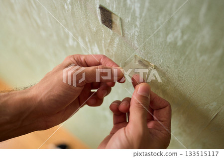 Closeup of textured plaster wall prep, Gently detaching thin tape from wall surface, Homeowner carefully removing adhesive strip to ready wall for repainting process Closeup of textured plaster wall prep, Gently detaching thin tape from wall surface, Homeowner carefully removing adhesive strip to ready wall for repainting process 133515117