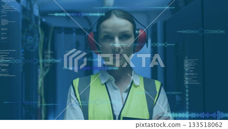 Monitoring tech in high-vis vest red earmuffs scanning racks in blue LED data center, code overlays 133518062
