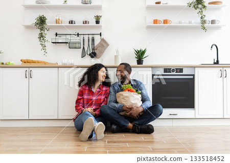 A smiling couple sits on the kitchen floor, looking at each other. The man holds a bag of groceries. A smiling couple sits on the kitchen floor, looking at each other. The man holds a bag of groceries. 133518452