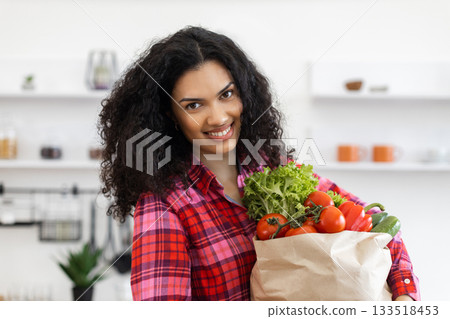 A smiling woman holds a paper bag filled with fresh vegetables in a kitchen setting. A smiling woman holds a paper bag filled with fresh vegetables in a kitchen setting. 133518453