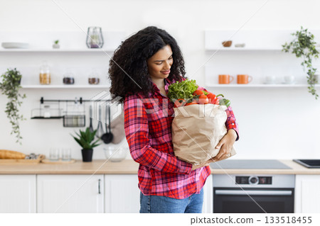A woman in a red plaid shirt carries a paper bag full of fresh groceries in a modern kitchen. A woman in a red plaid shirt carries a paper bag full of fresh groceries in a modern kitchen. 133518455