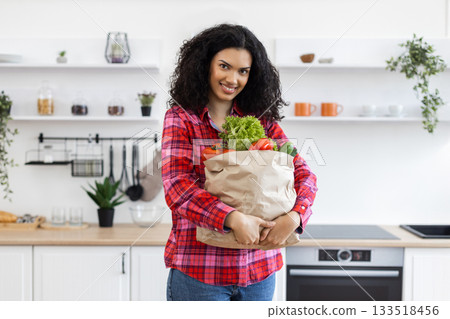 A smiling woman holds a paper bag filled with fresh produce in a bright, modern kitchen. A smiling woman holds a paper bag filled with fresh produce in a bright, modern kitchen. 133518456