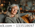A middle-aged woman is comfortably settled into a well-loved couch in her living room and listens to music through headphones. Precious moment of pause and personal indulgence 133518697