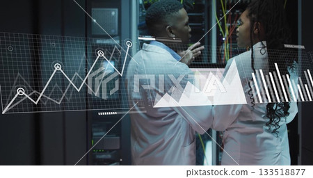 Gesturing engineers in lab coats reviewing metrics on laptop in data center, with racks and cables Gesturing engineers in lab coats reviewing metrics on laptop in data center, with racks and cables 133518877