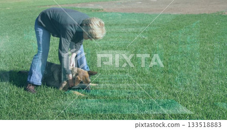 Bending senior woman holding brown-black dog collar on wide grassy field, with training session 133518883